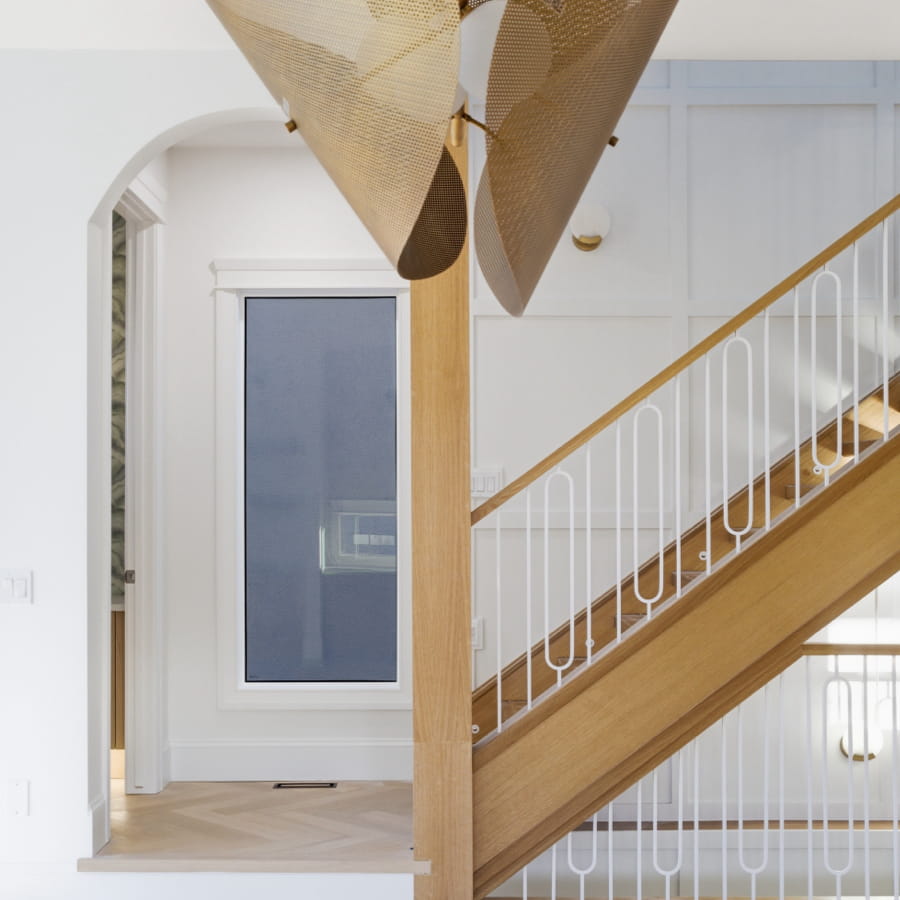 Modern staircase with wooden steps and white railings in a bright room. A large, gold light fixture hangs from the ceiling, adding elegance.