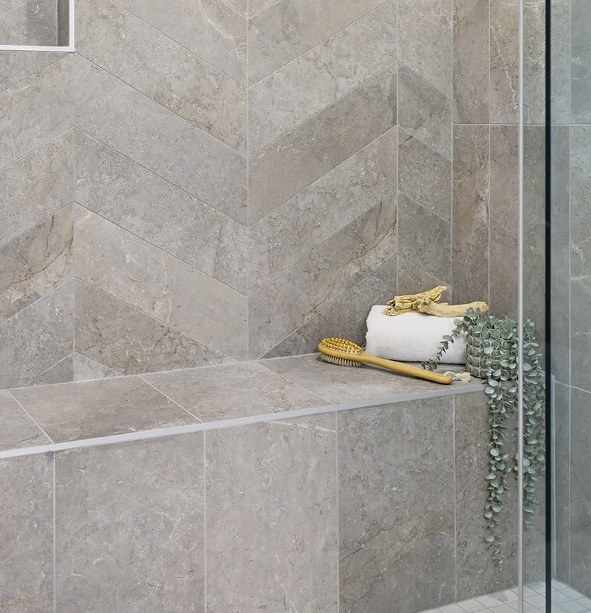Gray tiled shower with a built-in bench, rolled towel, wooden brush, and green leaves. Chevron pattern tiles add a sleek, modern feel.