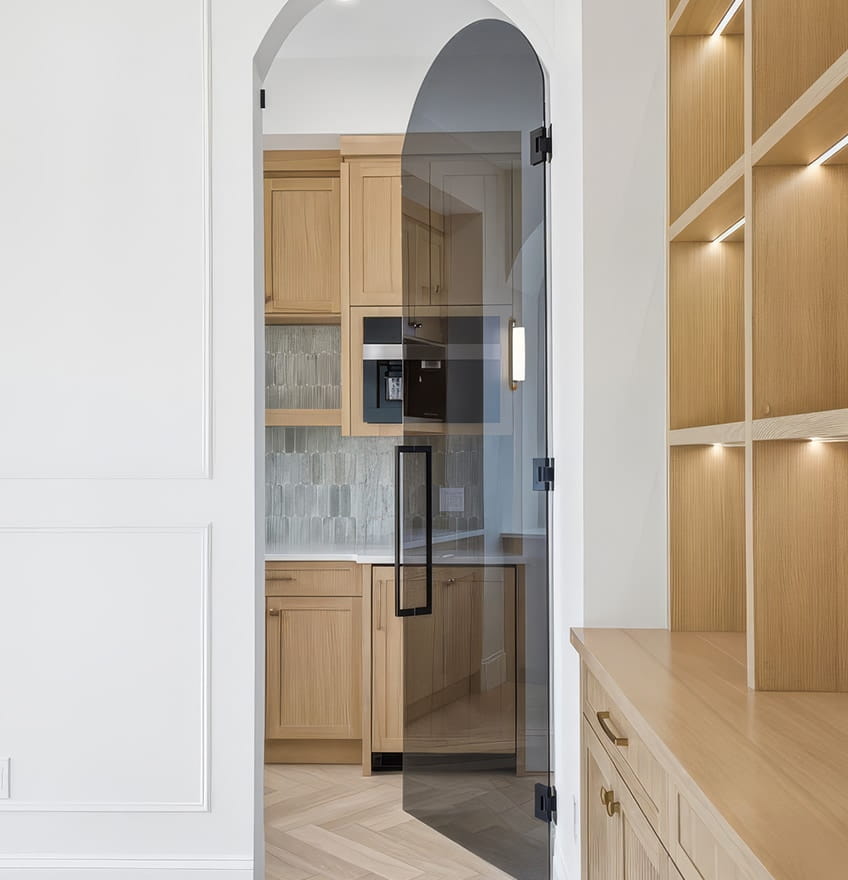 A modern kitchen with light wooden cabinets and a glass door with black hinges. The backsplash features gray tiles, and recessed shelving is lit softly.