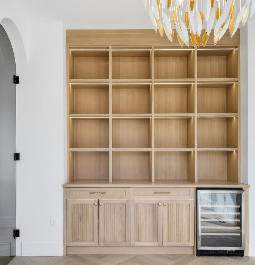 A modern kitchen with light wooden cabinets and a glass door with black hinges. The backsplash features gray tiles, and recessed shelving is lit softly.