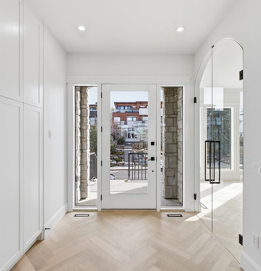 A modern kitchen with light wooden cabinets and a glass door with black hinges. The backsplash features gray tiles, and recessed shelving is lit softly.