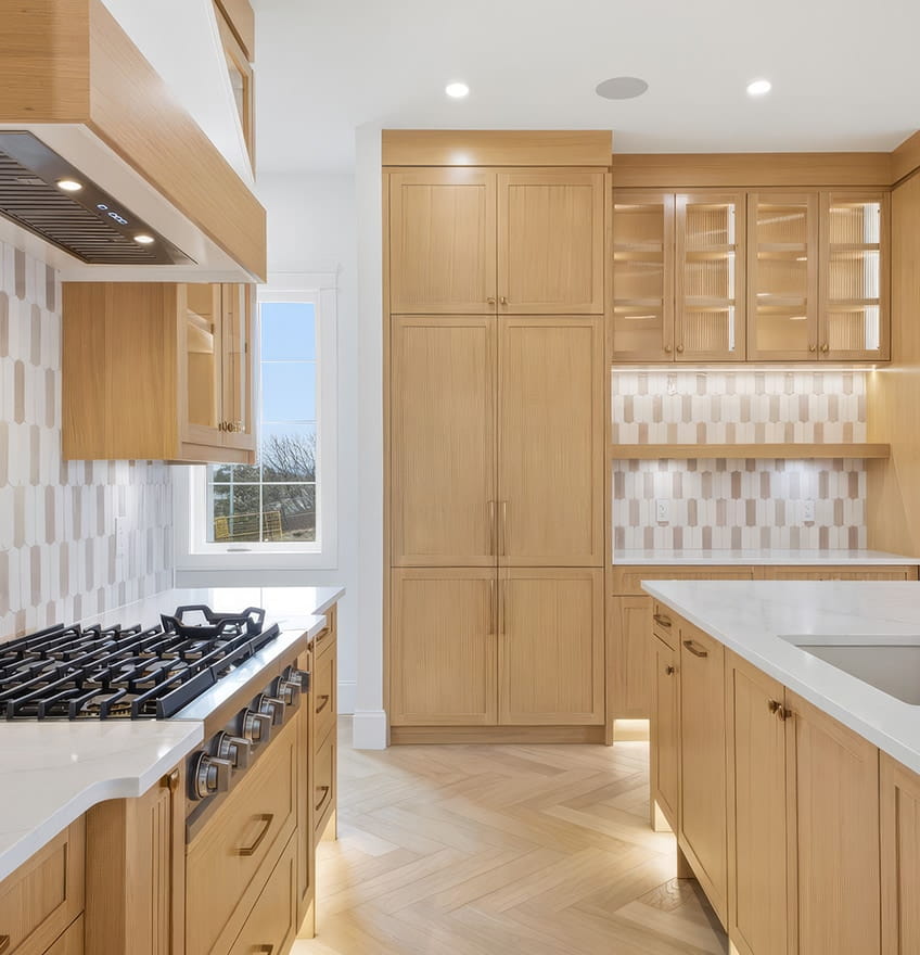 A modern kitchen with light wooden cabinets and a glass door with black hinges. The backsplash features gray tiles, and recessed shelving is lit softly.